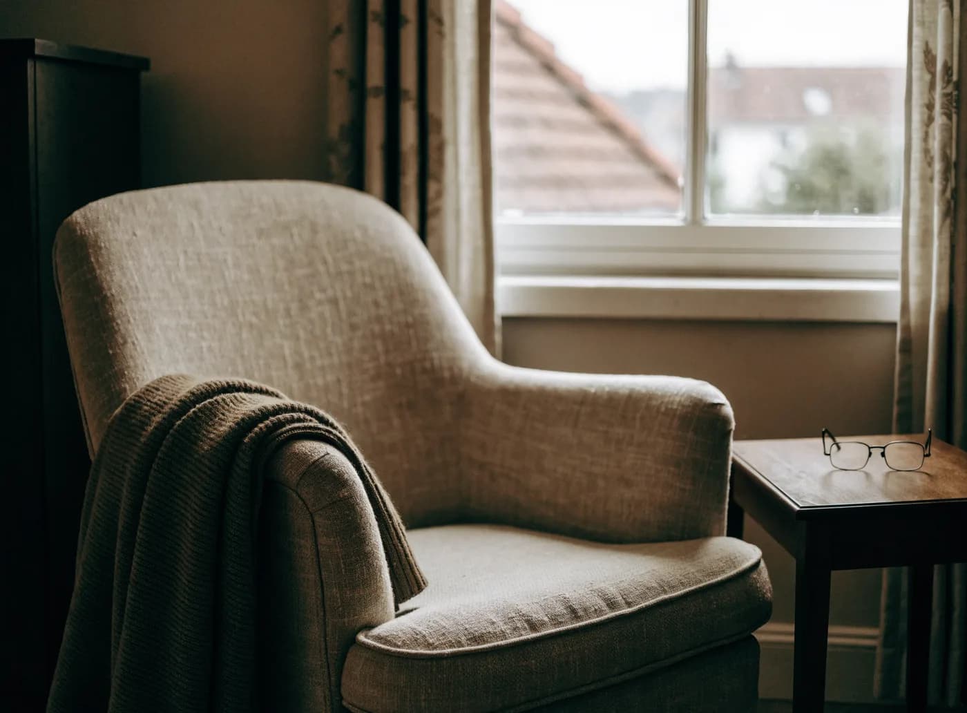 An empty armchair beside a window, a folded blanket over one arm and reading glasses on a side table